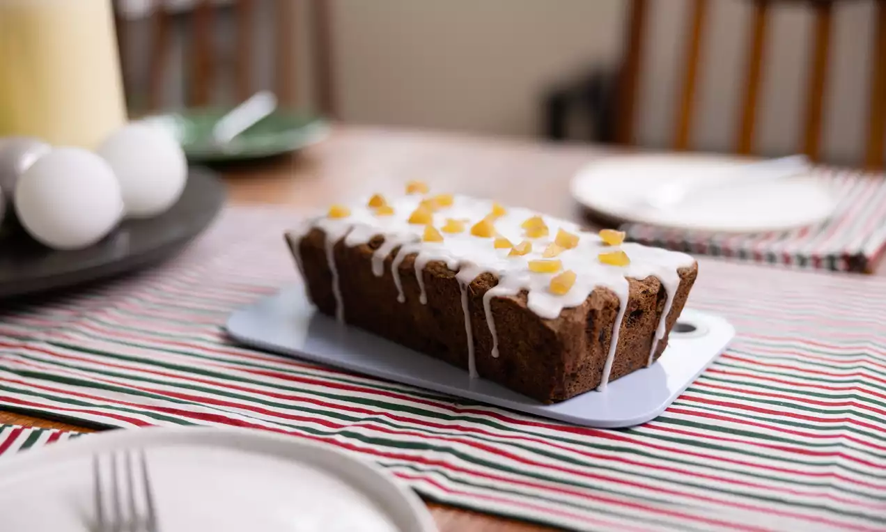 Chocolate chip fruit cake with a rich brown loaf shape, drizzled white icing and golden candied fruit pieces on a striped tablecloth